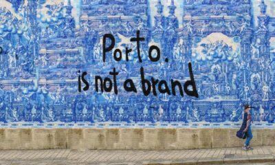 Capela das Almas, Porto, Portugal. Mural azul com desenhos detalhados de azulejos portugueses e a frase "Porto. is not a brand" escrita em preto no centro. No canto inferior direito, uma pessoa caminhando na calçada.