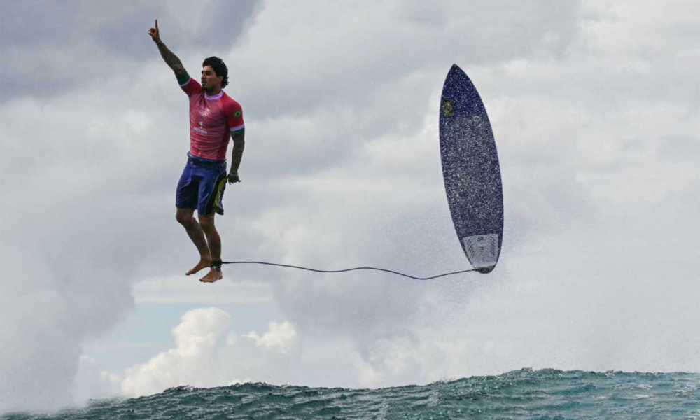 O surfista Gabriel Medina das Olimpíadas de Paris 2024. A prancha também está suspensa ao lado dele, com a correia presa ao tornozelo do surfista. Uma onda em movimento e um céu nublado compõem o cenário. A fotografia de Jérôme Brouillet.