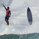 O surfista Gabriel Medina das Olimpíadas de Paris 2024. A prancha também está suspensa ao lado dele, com a correia presa ao tornozelo do surfista. Uma onda em movimento e um céu nublado compõem o cenário. A fotografia de Jérôme Brouillet.