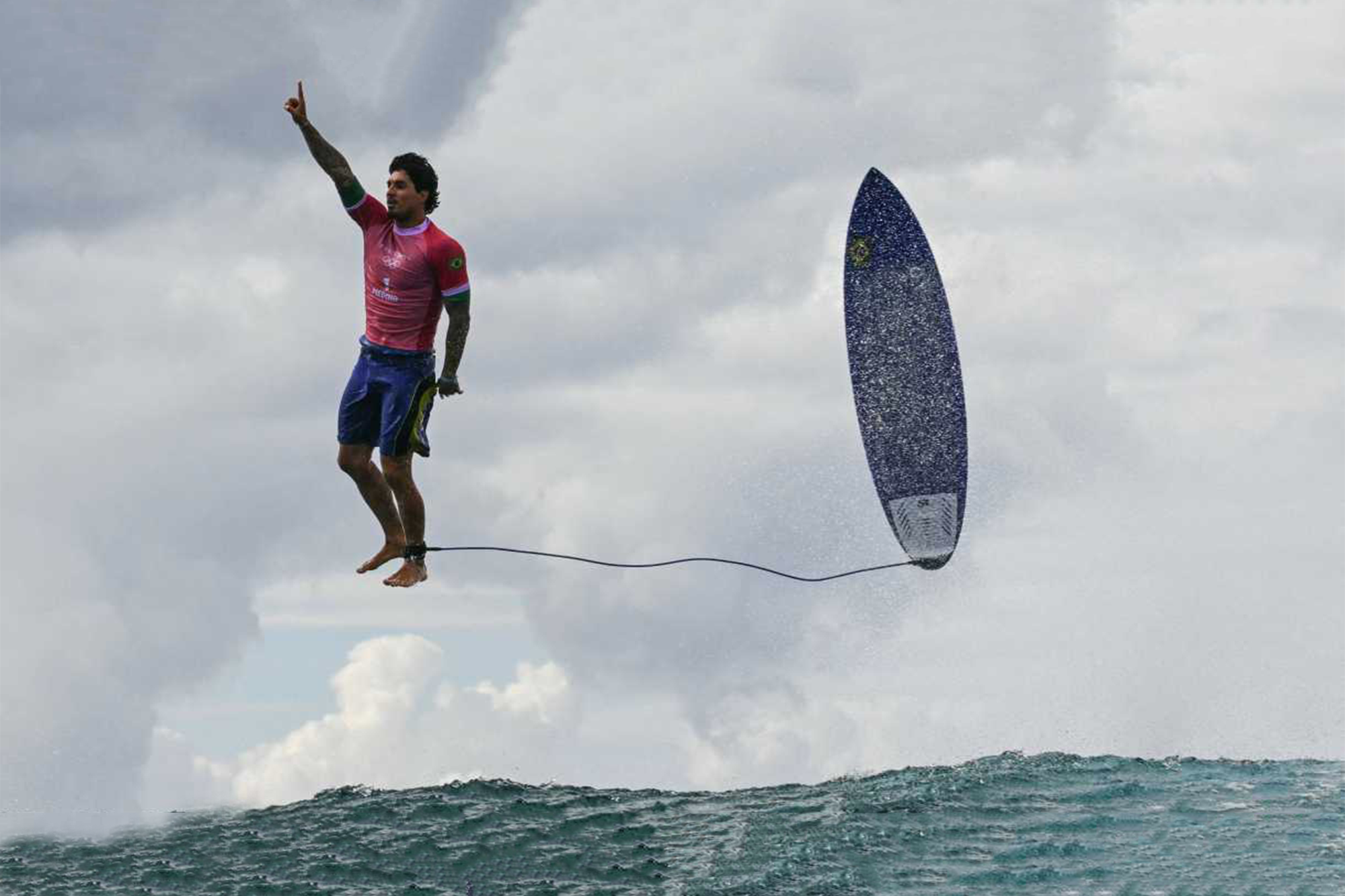 O surfista Gabriel Medina das Olimpíadas de Paris 2024. A prancha também está suspensa ao lado dele, com a correia presa ao tornozelo do surfista. Uma onda em movimento e um céu nublado compõem o cenário. A fotografia de Jérôme Brouillet.