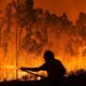 Morador tentando combater um dos focos de incêndio florestal durante a noite, em uma área densamente arborizada, envolta por chamas intensas. A silhueta da pessoa contrasta com o brilho alaranjado do fogo.